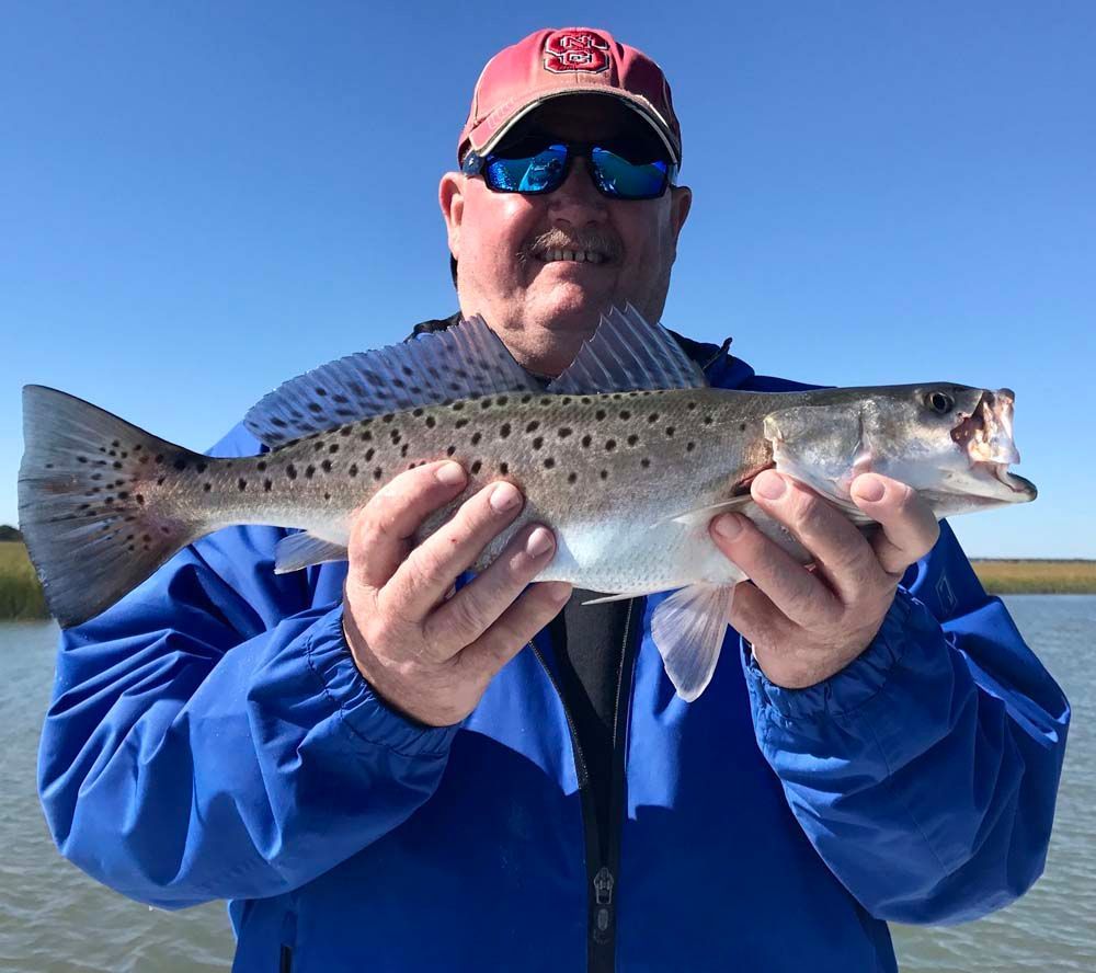 Man in blue jacket and cap holding a speckled fish outdoors under a clear blue sky
