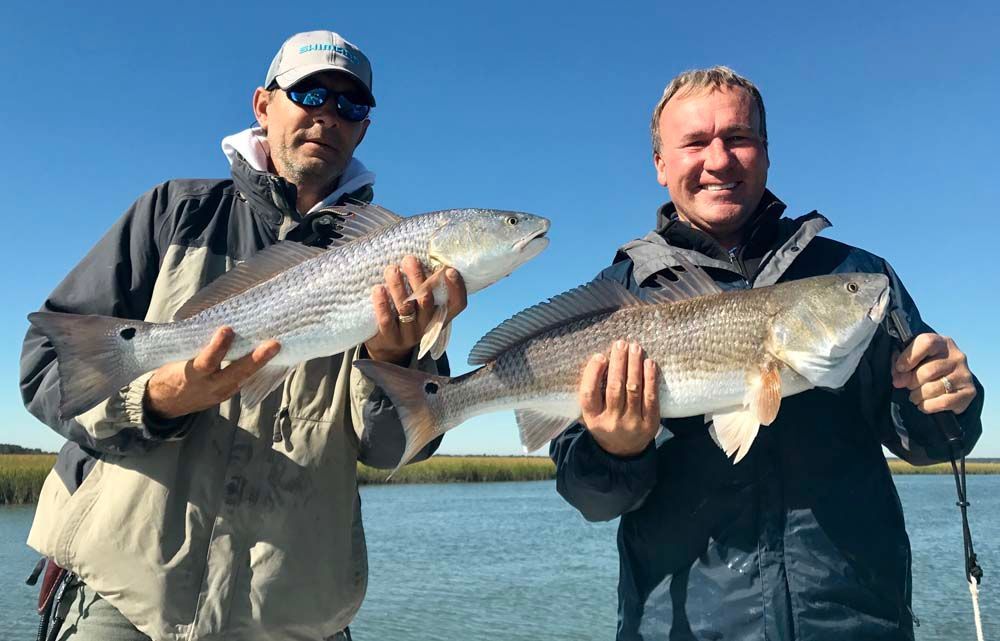 Two anglers holding large fish on a boat, with blue sky and water in the background