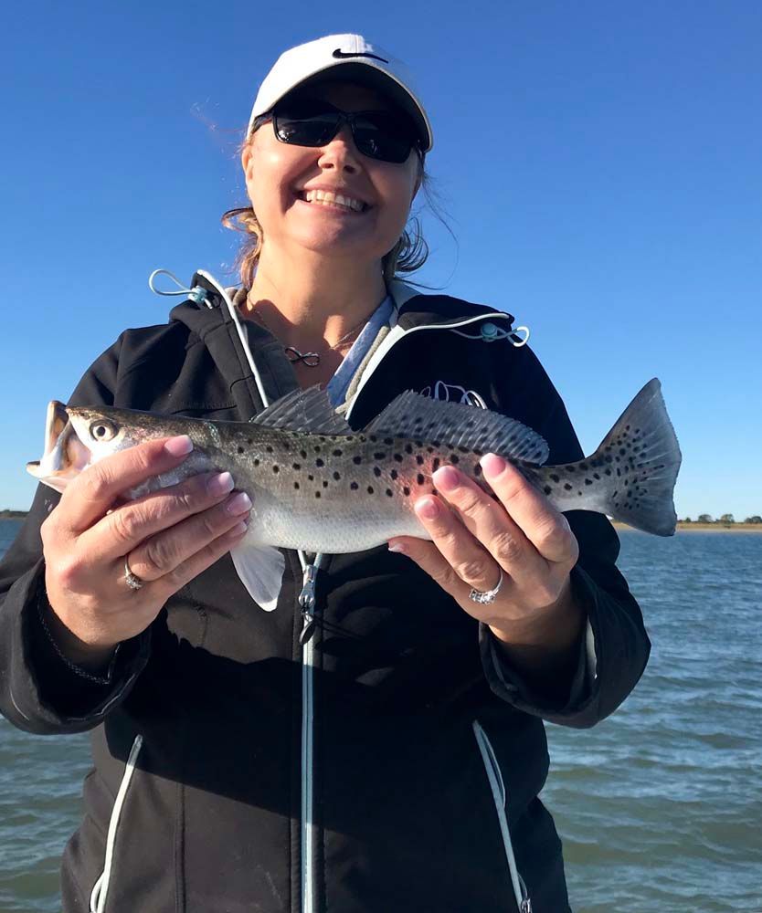 Woman holding a speckled trout on a boat, smiling under a sunny blue sky.