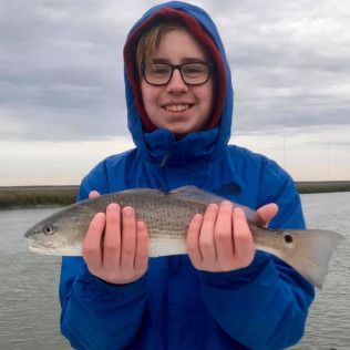 Person in blue jacket holding a fish at the water’s edge, smiling.