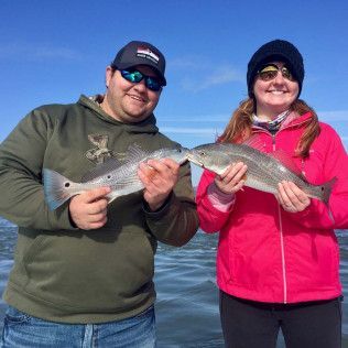 Two people smiling and holding a fish each by a lake on a sunny day