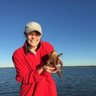 Person in a red jacket and cap holding an octopus by the water under a clear blue sky