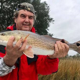 Man in a red jacket holding a large striped fish outdoors near marsh grass under a cloudy sky
