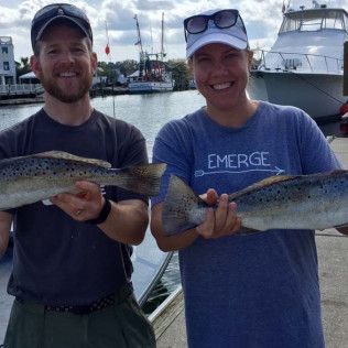 Two people holding fish on a dock in front of boats, smiling