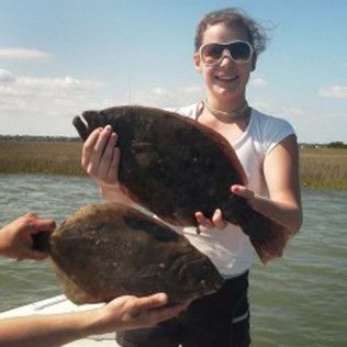 Person holding two large flat fish outdoors near the water, smiling in sunglasses