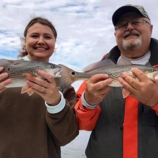 Two people smiling and holding fish outdoors under a cloudy sky