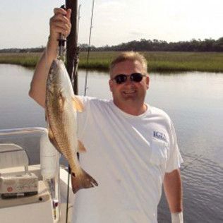 Man on a boat holding up a large fish by the water, wearing sunglasses and a white shirt.