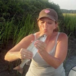 Person in a cap holding and kissing a fish outdoors near tall grass.