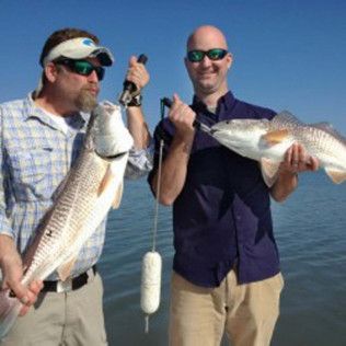 Two men holding large fish on a sunny boat, wearing sunglasses and casual clothes.