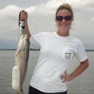 Person holding a large fish on a fishing line by the water, wearing sunglasses and a white T-shirt