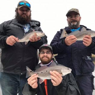 Three men in winter gear holding fish, posing outdoors in the snow.