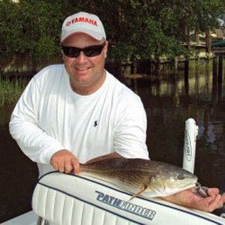 Man holding a large fish on a boat, wearing a white shirt, cap, and sunglasses by a calm wooded shoreline