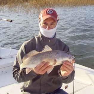 Person on a boat holding a fish over the water, wearing a cap and face covering