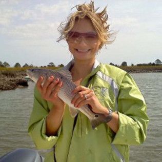 Person holding a fish near a river or marsh, wearing a light green jacket and sunglasses.
