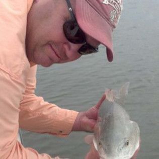 Person in a pink cap and sunglasses holding a fish over calm water.