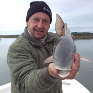 Man holding up a large silver fish on a boat with calm water and shore in the background