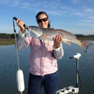 Person holding a large fish on a boat on calm water, wearing sunglasses and a pink vest.
