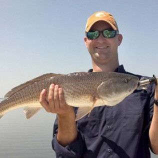 Man holding a large fish outdoors, wearing a yellow cap and sunglasses, smiling in bright sunlight