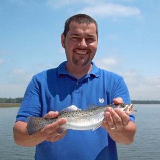 Man in blue polo smiling and holding a large fish by the water