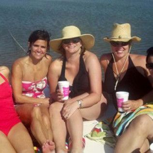 Three women in sun hats sit on a sandy beach, smiling with drinks in hand.