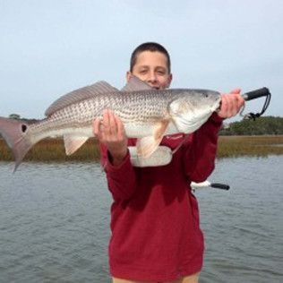 Person holding a large fish over water, wearing a red shirt with a cloudy sky in the background