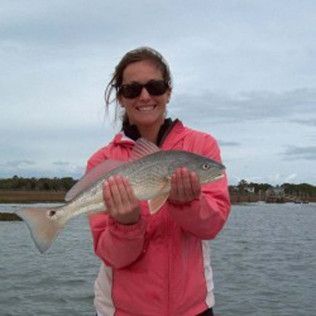 Person in a pink jacket and sunglasses holding a large fish by the water.