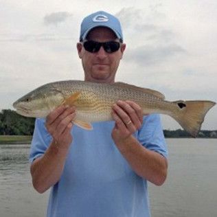 Man holding a large fish at the water’s edge, wearing a blue shirt, cap, and sunglasses.