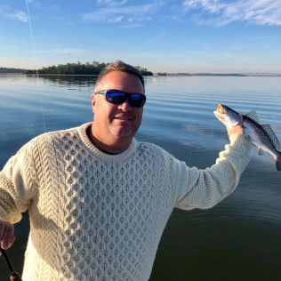 Man holding a fish in a calm lake at sunset, wearing sunglasses and a white sweater
