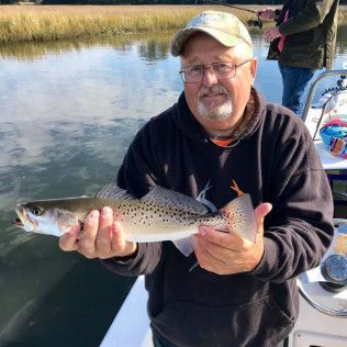 Man holding a large spotted fish on a boat beside calm water and marsh grasses