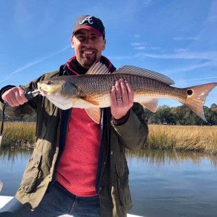 Person holding a large striped fish by a calm lake under a blue sky