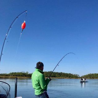 Person fishing from a dock with a red bobber on a calm blue river under a clear sky