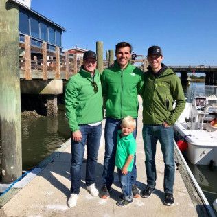 Four people in green jackets standing on a dock beside boats in bright sunlight