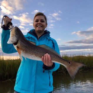 Person holding a large fish in a marsh at sunset, wearing a bright blue jacket