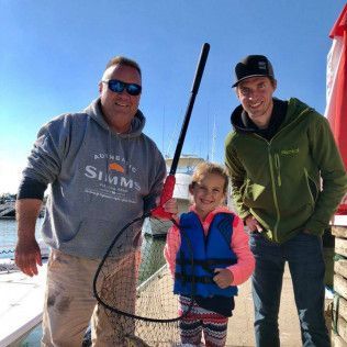 Three people smiling outdoors in winter gear beside a fishing net by the water