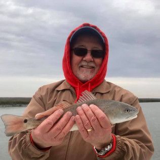 Person in red hood and sunglasses holding a fish by the water under cloudy skies