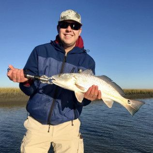 Man holding a large fish on a boat, wearing sunglasses and a blue jacket on calm water under a clear sky