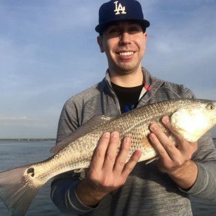 Man smiling while holding a large fish on a boat, wearing a Dodgers cap with water and sky behind him