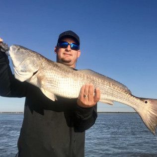 Person holding a large fish while standing on a boat or dock by the water, wearing sunglasses and a cap.