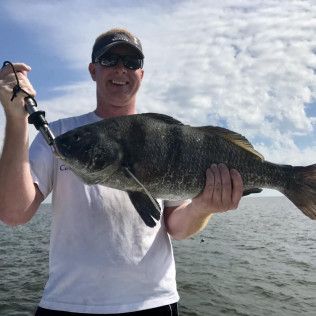 Person on a boat holding a large fish, wearing sunglasses and a cap, with open water and cloudy sky behind them