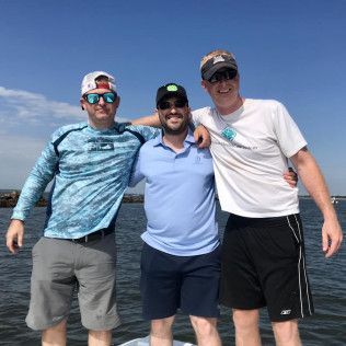 Three friends smiling with arms around each other on a sunny beach, wearing casual shirts and caps.