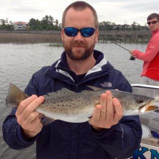Man in sunglasses holding a large fish on a boat, with another person fishing behind him.