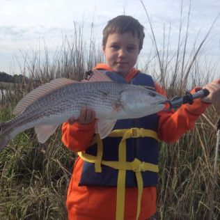 Boy in orange jacket and life vest holding a large fish in tall grass near water.