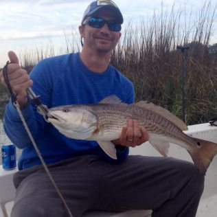 Man holding a large fish by a marsh, wearing a blue shirt and cap, smiling at the camera.