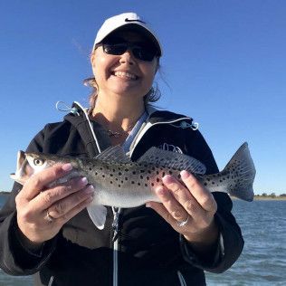 Smiling person in a cap holds a speckled fish at the beach with blue water and sky behind.