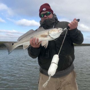 Angler holding a large silver fish on a boat or dock, wearing a red beanie and dark jacket under a cloudy sky