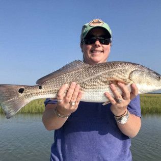 Person holding a large striped fish in front of a calm body of water under a clear blue sky
