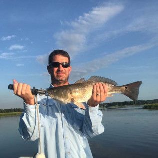 Man holding a large fish on a hook by a calm lake under a blue sky