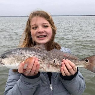 Young person smiling and holding a large fish at the water’s edge under an overcast sky