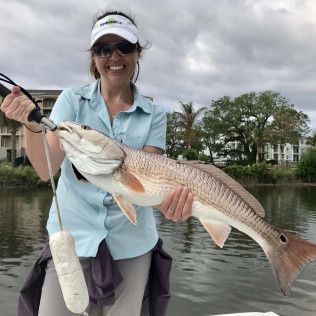 Person holding a large redfish on a boat by a calm river, wearing a visor and sunglasses.