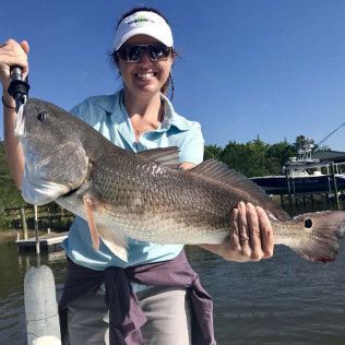 Person holding a large fish on a dock by the water, wearing a visor and sunglasses.